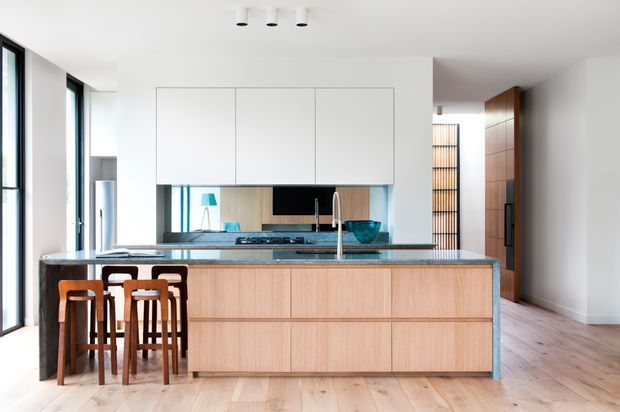 Bright contemporary kitchen with a large timber-fronted island, dark stone-look benchtop and integrated seating. White full-height upper cabinetry, a dark splashback, stainless-steel tapware and appliances, expansive black-framed glazing, and timber flooring define the space. A timber-lined hallway or adjoining area is visible beyond the kitchen.