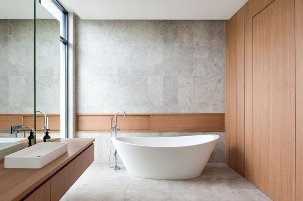 A refined contemporary bathroom featuring a freestanding white oval bathtub, wall-mounted and countertop basins, brushed metal tapware, warm timber cabinetry and wall panelling, large-format grey stone-look wall and floor surfaces, and a full-height mirror beside a narrow window. The room has a clean, calm, spa-like atmosphere with natural light and restrained detailing.