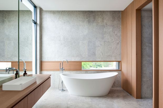 Contemporary bathroom featuring a freestanding white oval bathtub, wall-mounted white basin on timber vanity cabinetry, large grey stone-look wall and floor finishes, warm timber wall panelling and framing, and expansive glazing with a garden view. A large mirror reflects the vanity and basin, while vertical timber joinery frames an adjoining opening.