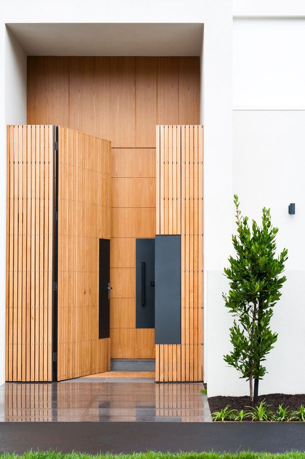Front exterior view of a contemporary residential entry. A tall recessed doorway is framed by warm vertical timber battens and timber wall panelling, with dark charcoal metal door panels and hardware. White rendered walls, polished stone or tile paving, and a narrow landscaped planter with a clipped tree complete the clean, minimalist facade.