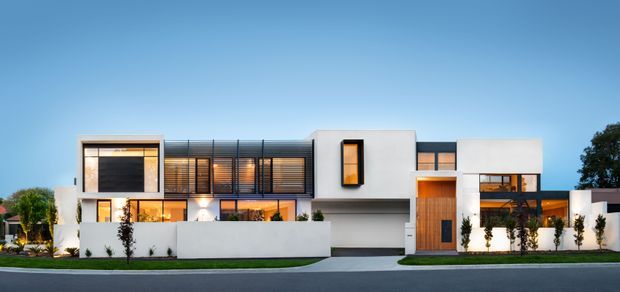 Wide exterior view of a large contemporary multi-level residence with a strongly rectilinear composition. The facade combines smooth white rendered volumes, extensive floor-to-ceiling glazing, dark horizontal louvres or screens, warm timber-toned entry and garage doors, and landscaped frontage enclosed by a low white wall. The house has a flat-roofed form and is photographed from the street at dusk or early evening with warm interior lighting visible.