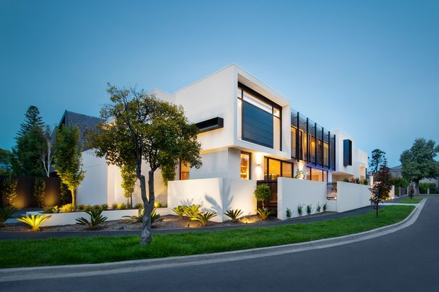A contemporary multi-level residential house viewed from the street at dusk. The home has crisp white rendered cubic forms, large dark-framed windows, projecting balconies or screens, and warm interior lighting. Low white boundary walls, structured planting, lawn, and a curved paved street define the landscaped frontage.