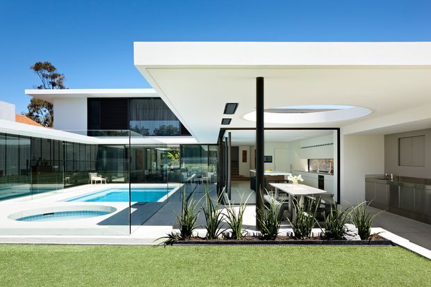 Bright exterior view of a contemporary flat-roof home organized around a swimming pool and lawn. The image shows strong horizontal roof overhangs, extensive floor-to-ceiling glazing with dark frames, a glass pool balustrade, a covered outdoor dining area, and an open-plan kitchen visible beneath the canopy. A circular roof opening/skylight and dark structural post add architectural interest.