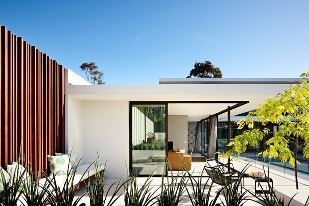 A contemporary single-storey residential facade and courtyard viewed from outside. The white flat-roofed building has expansive black-framed glazing and open sliding doors, with a tall vertical timber privacy screen along the left side. A paved courtyard with outdoor seating and planted landscaping is visible in front of the interior living space.