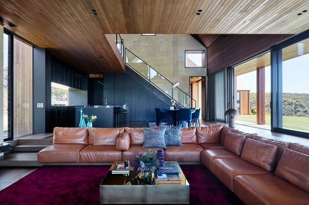 A spacious contemporary open-plan living area with a large cognac leather sectional sofa, deep burgundy rug, and low metallic coffee table. The room features a dramatic sloped timber-lined ceiling, dark vertical timber wall panelling, extensive floor-to-ceiling glazing, and a dining area beneath a visible stairway and mezzanine. The glazing opens toward a grassy, wooded landscape, while the interior combines warm timber, dark finishes, and rich leather furnishings.