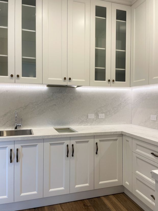 A compact L-shaped kitchen with white raised-panel cabinetry, a mix of glazed and solid upper cupboards, dark hardware, pale stone benchtops, and a matching full-height light grey veined splashback. Under-cabinet lighting illuminates the sink and preparation areas, with dark timber-look flooring below.