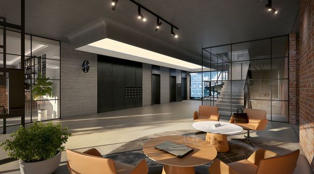 A contemporary commercial lobby or reception lounge with a double-height feel, dark ceiling, exposed brick feature wall, large black-framed glass partitions, dark elevator or service doors, and a relaxed seating arrangement around round timber and stone-topped tables. Track lighting and a large recessed ceiling light illuminate the space, while indoor planting and warm leather seating soften the industrial palette.