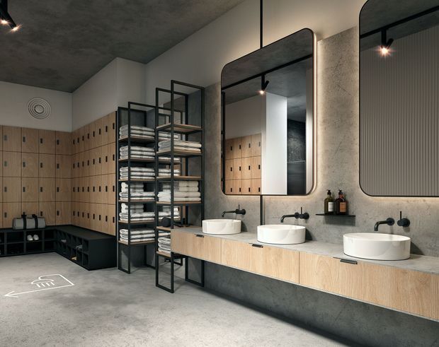 A contemporary communal bathroom or changing-room wash area with three round vessel basins on a long timber-look vanity, black wall-mounted taps, large rounded-rectangle mirrors, and open black metal shelving stacked with folded towels. The room has concrete-look walls, ceiling and floor, extensive light timber lockers along the back and side walls, black fixtures, and a utilitarian yet refined commercial or hospitality aesthetic.