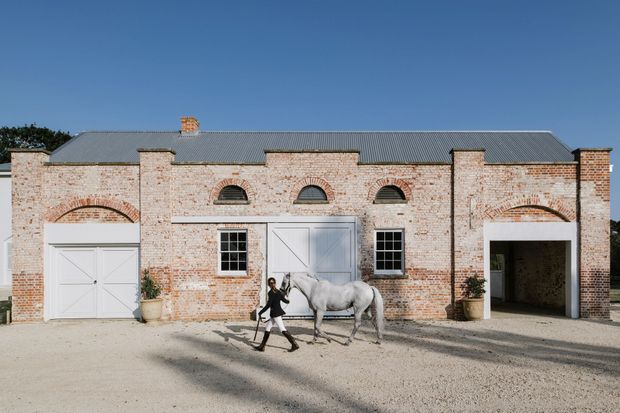 Front exterior view of a long, single-storey brick outbuilding or stable with a symmetrical facade, grey corrugated pitched roof, prominent brick pilasters, arched brick detailing, white doors and windows, and large garage-like openings. A horse and person are present in the forecourt, but the architecture is the primary subject.
