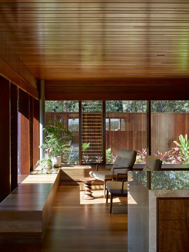A warm, richly timber-lined interior living area with a low built-in window seat or bench along the left wall, wood flooring, and extensive horizontal timber ceiling panelling. Floor-to-ceiling glazing opens the room to a planted outdoor setting, while upholstered lounge chairs and sculptural wooden stools create a quiet seating area. A partial foreground view shows a timber-fronted built-in cabinet or counter with a pale stone-like top. The space has strong natural light, deep timber tones, and an indoor-outdoor connection.