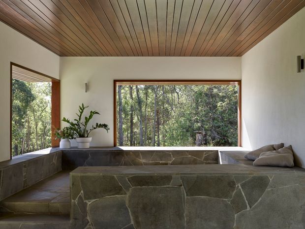 A serene, nature-facing interior lounge with extensive built-in stone seating and a low stone-clad platform. Large timber-framed windows overlook dense woodland, while a warm timber-lined ceiling spans the room. White plaster walls, restrained black wall lights, and potted greenery create a calm, organic atmosphere.