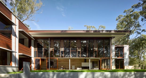 Exterior view of a contemporary multi-level residence set among mature trees. The building features extensive floor-to-ceiling glazing, warm timber wall cladding and screens, dark metal framing, strong horizontal roof planes, and a sheltered ground-level outdoor living area opening onto a lawn.