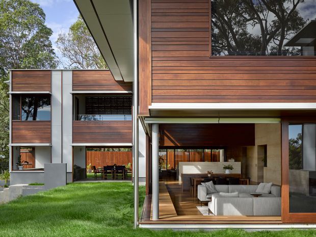 Modern multi-level residential house exterior with strong indoor-outdoor connections. The facade combines horizontal warm timber cladding, white rendered wall panels, extensive glazing, deep roof overhangs, and large sliding glass openings. Through the glazing, a contemporary living room with a light sectional sofa and timber flooring is visible, while a dining area and outdoor lawn occupy the foreground and surrounding site.