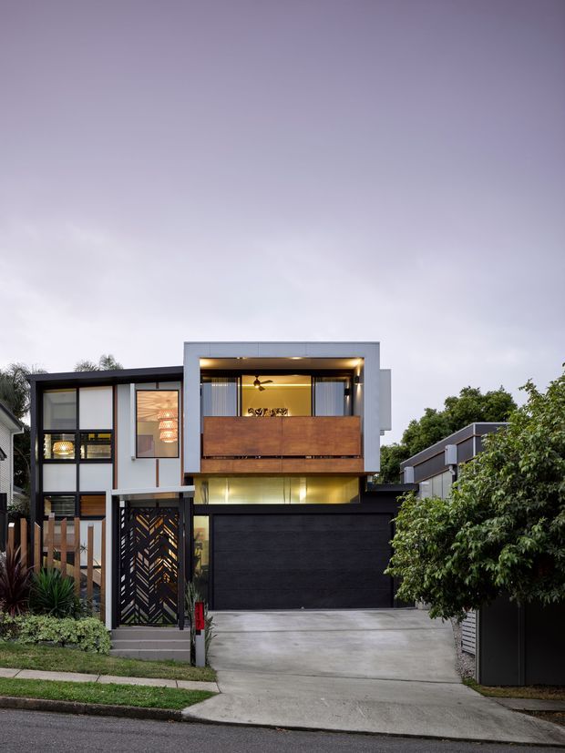 Street-facing view of a contemporary multi-level residence with a strong geometric facade. The house combines white rendered wall planes, dark framed glazing, a projecting upper-level box, warm timber screening, and a large dark garage door. A patterned entry gate, landscaped planting, concrete driveway, and neighbouring urban homes are visible.
