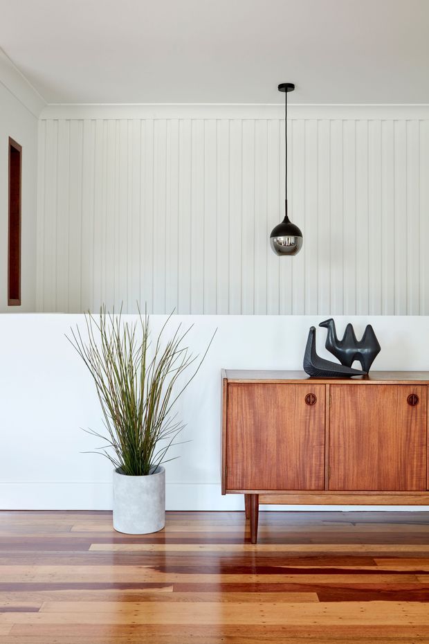 A refined interior vignette featuring a mid-century-style timber sideboard against a white vertically panelled wall. The room has polished timber flooring, a minimalist black pendant light, a tall potted grass plant, and a sculptural black decorative object on the sideboard. A narrow framed artwork or inset is visible at the left edge.