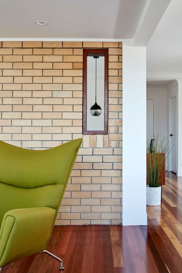 Interior view of a bright residential living area featuring an exposed pale brick feature wall, polished dark timber flooring, and a vivid olive-green upholstered armchair in the foreground. A narrow timber-framed wall mirror reflects a hanging black pendant light, while a white structural column opens toward a hallway with additional timber flooring and a tall potted plant.