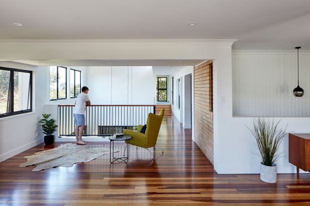 Bright open-plan residential interior showing a raised living area and hallway with polished timber flooring, white walls and ceilings, extensive black-framed glazing, a timber balustrade, and a prominent vertical timber feature wall. A mustard lounge chair, small side table, indoor plants and a partial timber console furnish the space.