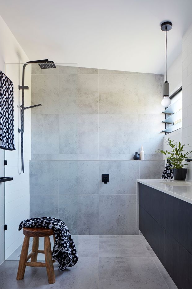 A bright contemporary bathroom featuring a walk-in shower with a black overhead and handheld shower system, large pale grey stone-look wall and floor tiles, a floating dark vanity with a light benchtop, white wall tiles, black-framed louvred windows, and a suspended pendant light. Monochrome patterned textiles and a timber stool add contrast and warmth.