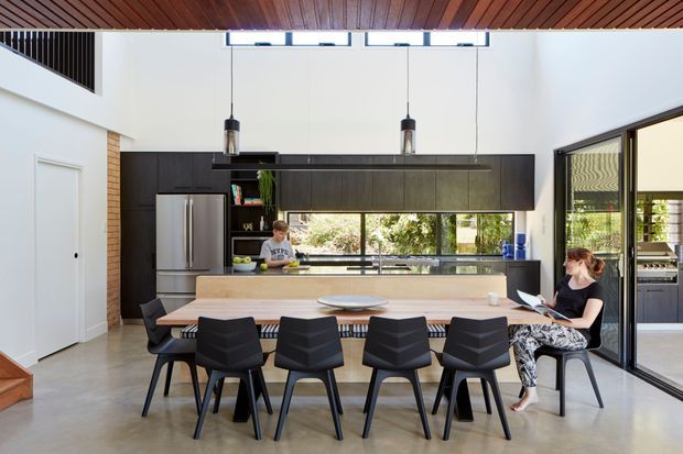 A bright, open-plan contemporary kitchen and dining area with a long light-toned island and integrated dining table surrounded by black chairs. Dark timber cabinetry spans the rear wall, with stainless-steel refrigeration, a polished dark benchtop, pendant lights, clerestory glazing, and large glazed doors. The room has a polished concrete floor, exposed brick detail, and a warm timber slatted ceiling.