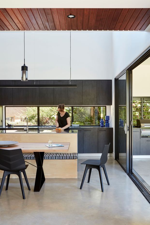 Contemporary kitchen and dining area with dark timber cabinetry, a pale island bench, polished concrete flooring, and a warm timber-lined ceiling. Full-height glazing and sliding glass doors connect the kitchen to an outdoor garden setting. Black pendant lights, dark dining chairs, and a built-in bench seat complete the clean, modern interior.