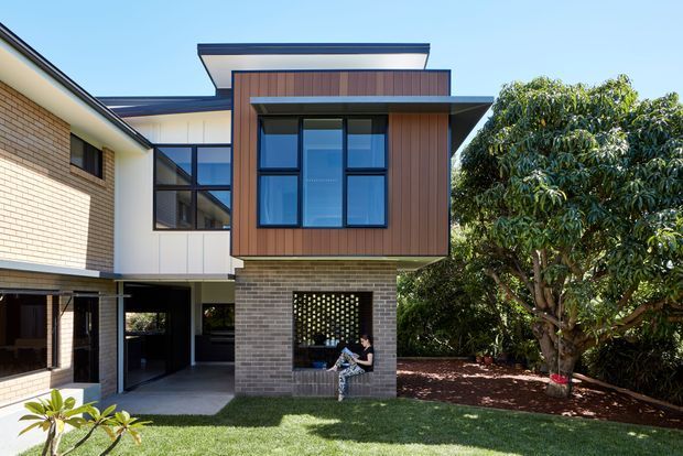 Exterior view of a contemporary multi-level residence with a flat-roofed projecting upper volume clad in dark timber, large black-framed windows, and a pale brick lower level. The facade includes a sheltered open terrace or seating area beneath the upper floor, with landscaped garden and lawn in the foreground.