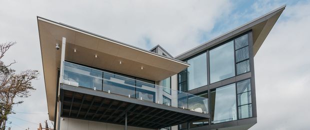 Exterior view of a contemporary multi-level residence with dramatic cantilevered roof planes, extensive floor-to-ceiling glazing, glass balcony balustrades, and a dark structural undercroft. The building has a sculptural, angular form and is photographed from below against a cloudy sky.