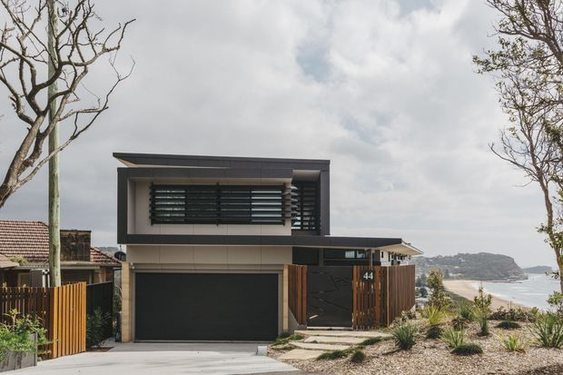 Contemporary two-storey coastal residence viewed from the street, with a flat projecting roof form, dark horizontal louvres, pale rendered upper walls, timber privacy screens, and a dark garage door. A sandy shoreline and headland are visible beyond the property, with drought-tolerant landscaping and mature trees framing the image.
