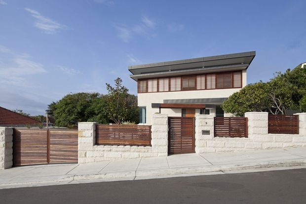 Street-facing view of a contemporary two-storey residence with a broad low-pitched roof, white rendered walls, upper-level timber screening, and a front boundary combining white masonry walls with horizontal dark timber slat fencing and matching pedestrian and vehicle gates. The house sits along a paved suburban street with established planting.