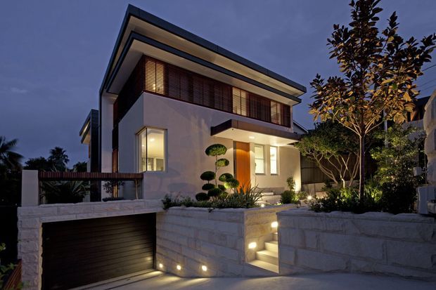 Twilight exterior view of a refined two-storey contemporary home on a sloping site. The house has clean rendered walls, strong horizontal roof and balcony lines, extensive upper-level glazing with dark timber or wood-look louvres, a warm timber entry door, landscaped planting, an illuminated stairway, and an integrated dark garage door set into a pale masonry retaining wall.