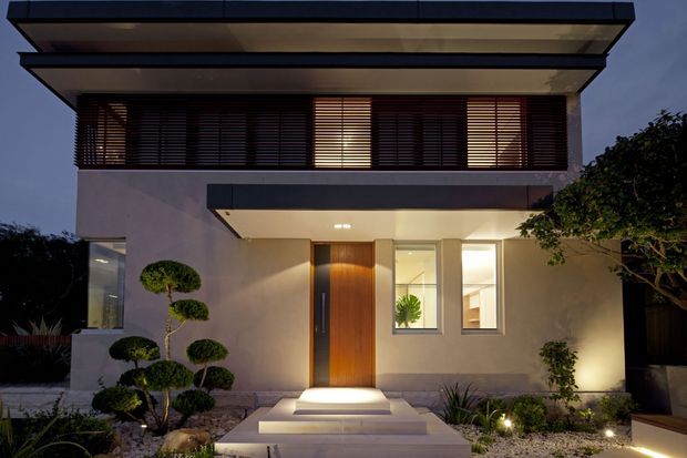 Night-time frontal view of a refined modern two-storey house facade. The home features a strongly projecting flat roof with layered horizontal planes, a broad upper-level louvred screen, smooth pale rendered walls, dark trim, and a tall warm timber entry door beneath a covered porch. Illuminated steps lead through a minimalist landscaped forecourt with sculpted topiary and gravel planting.