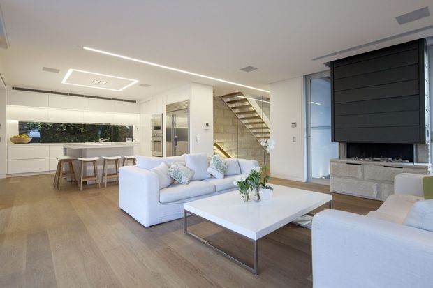Bright open-plan contemporary living space with a white kitchen and island dining bench at the rear, a large pale sectional sofa and armchair around a low rectangular coffee table, and a modern black feature fireplace wall. A glazed stairway with timber treads and a glass balustrade rises behind the seating area. The room has light timber flooring, white walls and ceiling, extensive glazing, and integrated linear lighting.