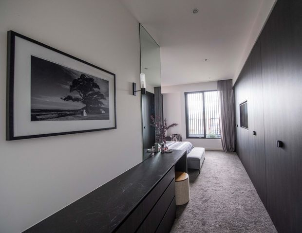A narrow, contemporary living room or lounge with a long dark built-in console and drawers, dark vertically textured wall panelling, a wall-mounted television, and a compact sofa facing the TV. A large black-and-white landscape artwork hangs on the pale wall, while full-height curtains and vertical window blinds bring filtered daylight into the room. The palette is restrained in charcoal, grey, white, and muted timber tones, creating a refined, calm atmosphere.