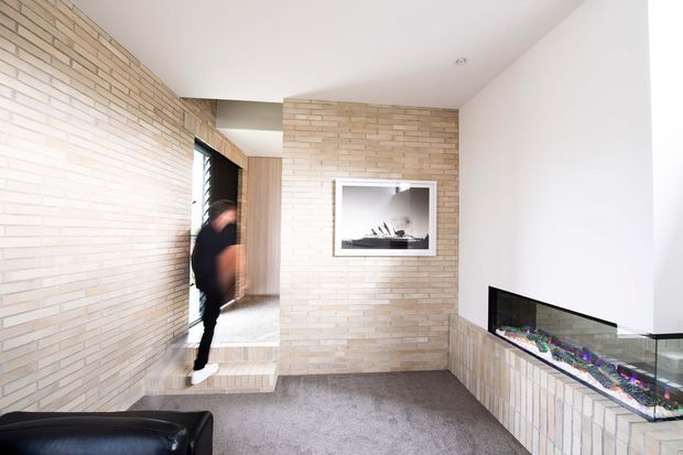 Bright contemporary interior hallway or living area featuring extensive pale brick wall finishes, a carpeted floor, recessed ceiling lighting, timber detailing around a doorway, and a long built-in linear fireplace with a brick-clad surround. A framed artwork is mounted above the central brick wall, and part of a dark leather sofa is visible in the foreground.