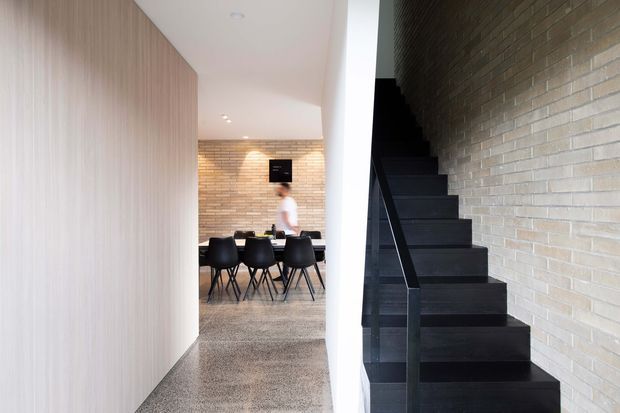 A contemporary residential interior viewed from a hallway, with a dark open staircase running beside a pale exposed-brick wall. Beyond the passage is a dining area with a dark rectangular table and black chairs, while the left wall is clad in light timber panelling. The space is bright, clean, and minimalist with strong material contrast between the timber, brick, white surfaces, and black stair.