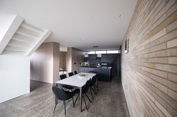 A contemporary open-plan dining and kitchen area beneath a white staircase. The room features a long light-coloured dining table with dark chairs, dark vertically ribbed kitchen cabinetry and island, warm timber-toned joinery, a full-height exposed light brick wall, recessed ceiling lighting, and a carpeted floor.
