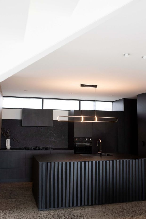 A contemporary dark-toned kitchen featuring a large island with vertically fluted cabinetry, integrated dark cabinetry, a black stone-look splashback, built-in oven, gas cooktop, and curved black tap. High horizontal clerestory glazing brings in diffuse daylight, while a linear pendant light hangs over the island. The space has a restrained, refined monochromatic palette with warm lighting and a polished modern feel.