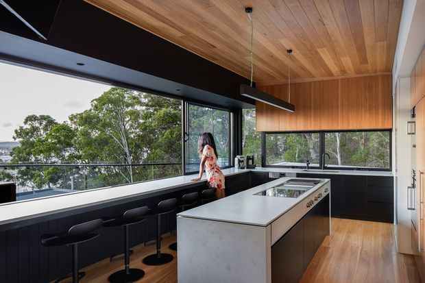 Contemporary kitchen with a large central island, dark lower cabinetry, pale stone benchtop, extensive glazing, and warm timber lining across the ceiling and upper cabinetry. A breakfast bar with black stools runs along the window, overlooking leafy surroundings and a distant water view. A linear pendant light hangs above the island.