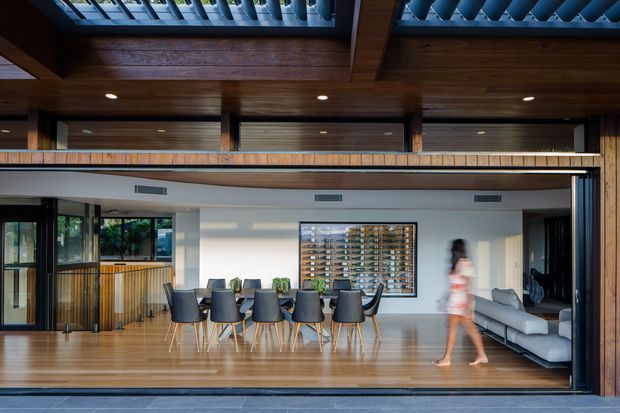 Open-plan contemporary living and dining area viewed through a wide glazed opening, with a long timber dining table, upholstered dining chairs, a grey sofa, extensive timber ceiling beams and lining, polished timber flooring, and a mezzanine-like upper level. Large operable glazing connects the interior to the exterior, while a skylight or roof opening and exposed structural timber create a strong indoor–outdoor architectural character.