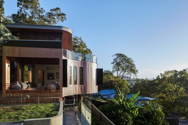 Exterior view of a contemporary multi-level home set on a steep, leafy suburban site. The building features curved forms, dark timber cladding, extensive glazing, projecting balconies, and a flat roof, with glimpses of an outdoor living area and surrounding houses beyond.