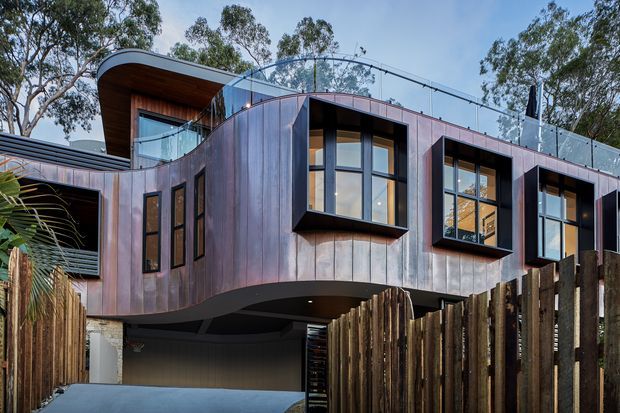 Exterior view of a contemporary multi-level residence with a sculptural, curved facade clad in vertically oriented dark timber-toned panels. Large black-framed windows project outward from the facade, while a rooftop terrace with clear glass balustrades is visible above. The home is elevated over a recessed driveway or carport and surrounded by mature trees and timber fencing.