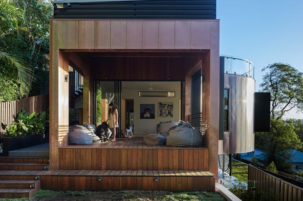 Exterior view of a compact contemporary timber-clad pavilion or extension set into a lush sloping site. The open front reveals a relaxed lounge area with beanbag seating, timber decking, dark-framed glazing, and a small interior room beyond. A cylindrical metal-clad structure projects from the side, while stairs, balustrades, planted landscaping, and neighbouring treetops reinforce the elevated hillside setting.