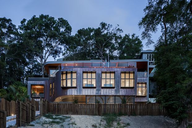 Twilight exterior view of a contemporary multi-level residence set among mature trees. The building features a sculptural curved upper roofline, extensive warm-lit grid windows, vertically finished grey cladding, dark timber fencing, and a partially visible elevated upper volume.