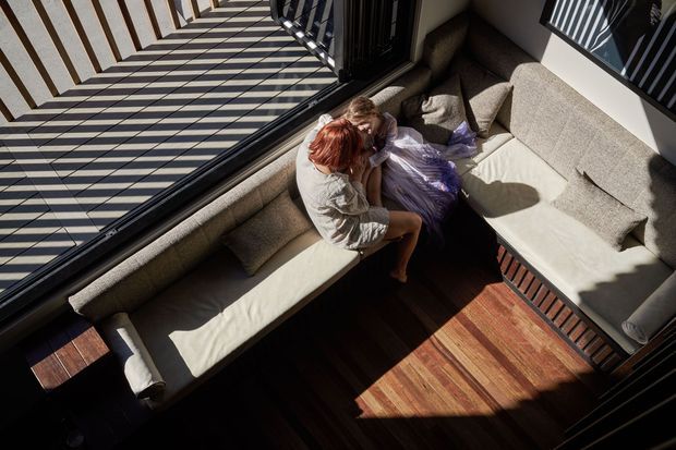 High-angle view of a warm, light-filled living room with a large L-shaped upholstered sectional sofa beside floor-to-ceiling glazing. Strong linear shadows from the exterior balustrade fall across the dark timber floor and seating. Two people are seated on the sofa, while the visible architecture is otherwise restrained and contemporary.