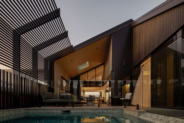 A dramatic contemporary residential courtyard viewed from beside a swimming pool. The home features dark vertical cladding, extensive glazing, warm timber-lined interiors, and a large covered pergola with closely spaced slatted beams. A lounge area sits beside the pool, while the glazed central volume reveals a warm, open-plan interior and a skylight.