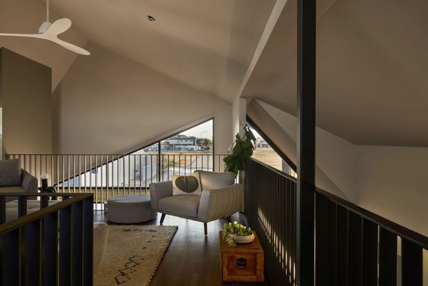 A contemporary loft-like living area on an upper landing, with a sloped ceiling, angular floor-to-ceiling glazing, dark metal balustrades, and warm timber flooring. The space contains a pale sofa, round ottoman, textured rug, indoor plant, and timber side table, with a stair or void visible in the foreground and residential buildings seen through the windows.