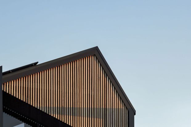 Close-up exterior view of a contemporary gabled building facade, featuring a dark structural frame and closely spaced vertical timber-look slats across the triangular gable end. The clear sky forms the background, emphasizing the roofline and rhythmic screen facade.