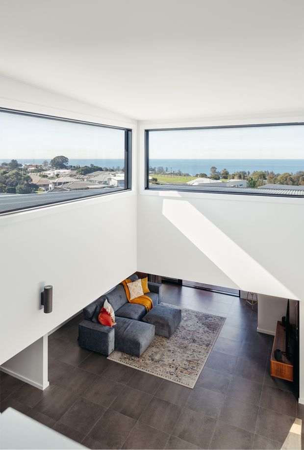 A bright, contemporary double-height living room with expansive corner windows overlooking a coastal residential landscape and ocean. The space features white walls, dark tiled flooring, a charcoal sectional sofa with colourful cushions, a patterned area rug, and strong natural sunlight creating geometric shadows. The elevated viewpoint highlights the open void and generous glazing.