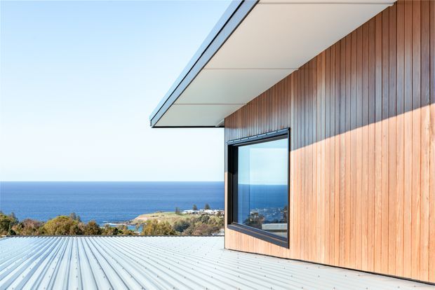 Exterior architectural view of a contemporary coastal home, featuring vertical timber wall cladding, a broad projecting soffit, dark-framed glazing, and a corrugated metal roof in the foreground. The ocean and coastal landscape are visible beyond the building.