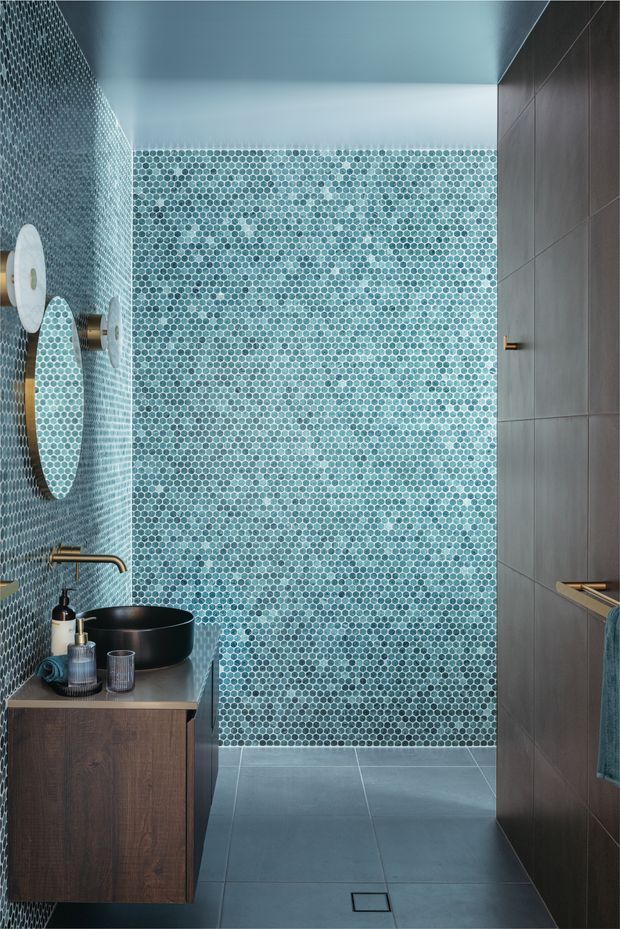 A narrow contemporary bathroom with a floating dark timber vanity, matte black vessel basin, wall-mounted brass tapware, and a large round mirror. The rear and left walls are covered in small turquoise and pale blue mosaic tiles, while the floor and right wall use large cool grey tiles. Brass hardware and a teal towel add warm and cool accents.