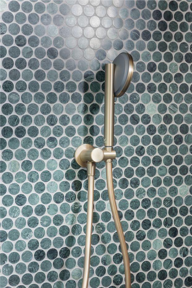 Close-up of a contemporary shower fixture mounted against a wall covered in small round green and grey-blue mosaic tiles. The shower rail, hand shower, hose, and controls have a brushed brass finish, creating a refined spa-like bathroom detail.
